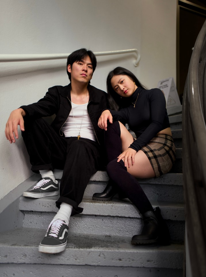 A portrait of two individuals sitting on stairs, with one leaning on the other. The individuals appear to be posing in a studio-like environment.