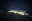 A nighttime landscape shows a mountain range with illuminated slopes and a cloudy sky. The scene is illuminated by lights scattered across the mountain. blurred out low res