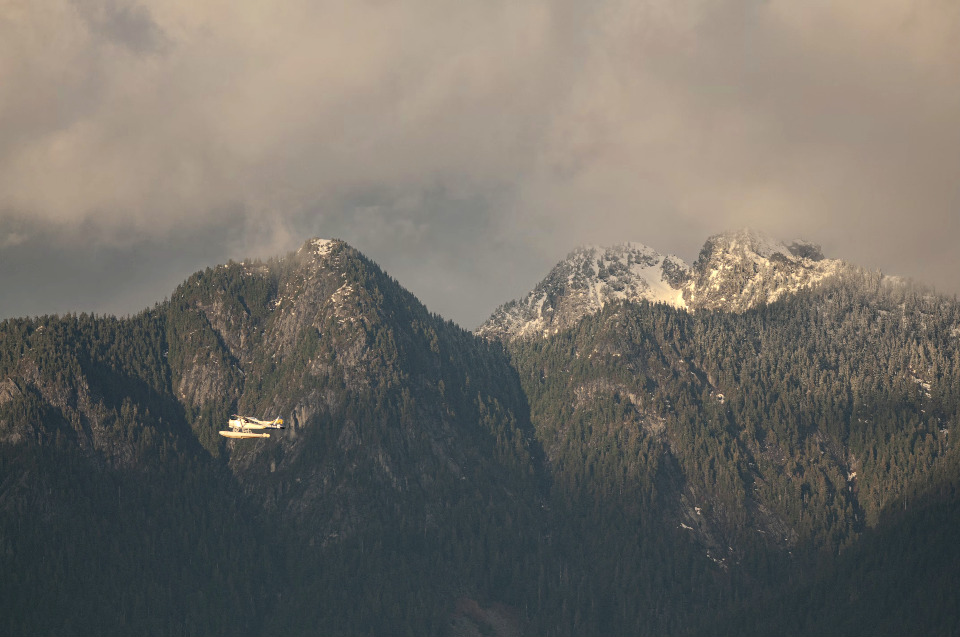 A seaplane soars against a backdrop of towering, snow-dusted mountains. The scene is enveloped in the soft light of a cloudy day.